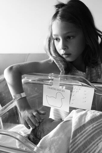 The eldest sister has her hand in the hospital bassinet, touching her head while gazing to the left. She has a neutral expression.