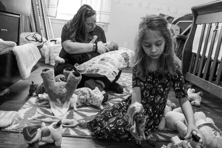 An occupied mother lays her infant on a pillow. The eldest sister is playing with a doll. The room surrounding them has toys and laundry strewn about.
